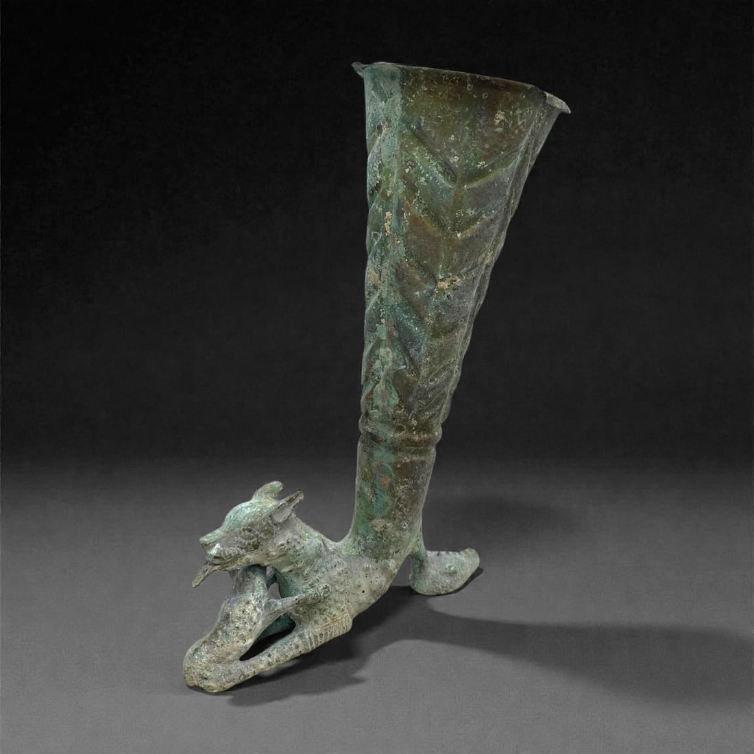 An Ancient Near Eastern Bronze Rhyton with Feline Base, 272g, 43AA: A ritual bronze rhyton, measuring 22 cm in height and weighing 272g, featuring a tapering, flared bowl with incised geometric decoration, supported by a dynamic feline base. The feline is depicted in
