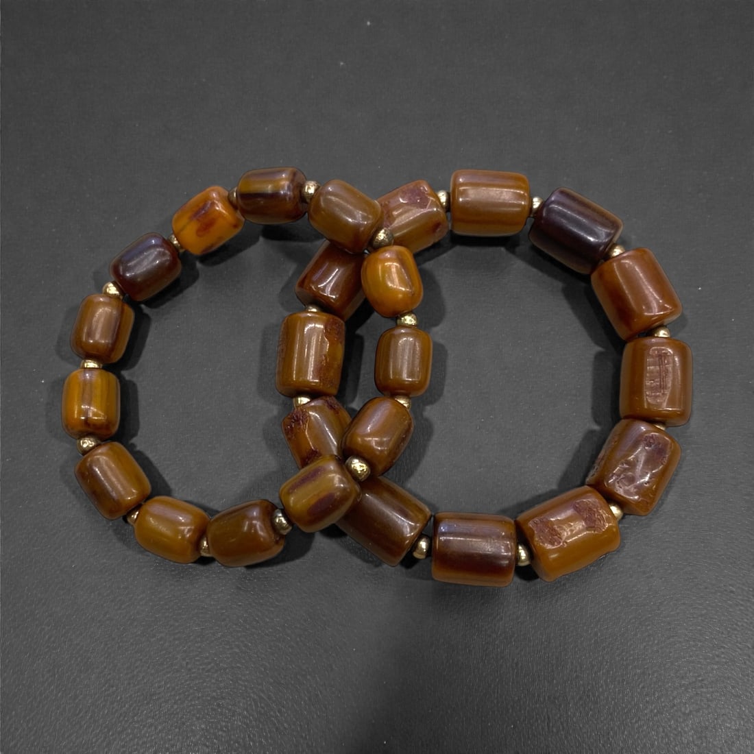 A Group of Two Amber Beaded Bracelets, 33g, 90AA: Comprising two stretch bracelets composed of polished barrel-shaped amber beads, alternating with small gold-toned spacer beads. The amber exhibits a rich, translucent reddish-brown to honey hue, with