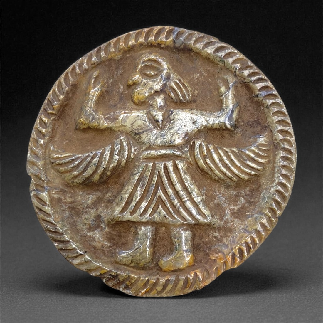 An Ancient Near Eastern Stone Plaque, 45g, 88AA: Carved from a single piece of stone with a mottled, earthy brown and yellowish matrix, the plaque is circular in form. The central field is raised in relief, depicting a standing human figure with out