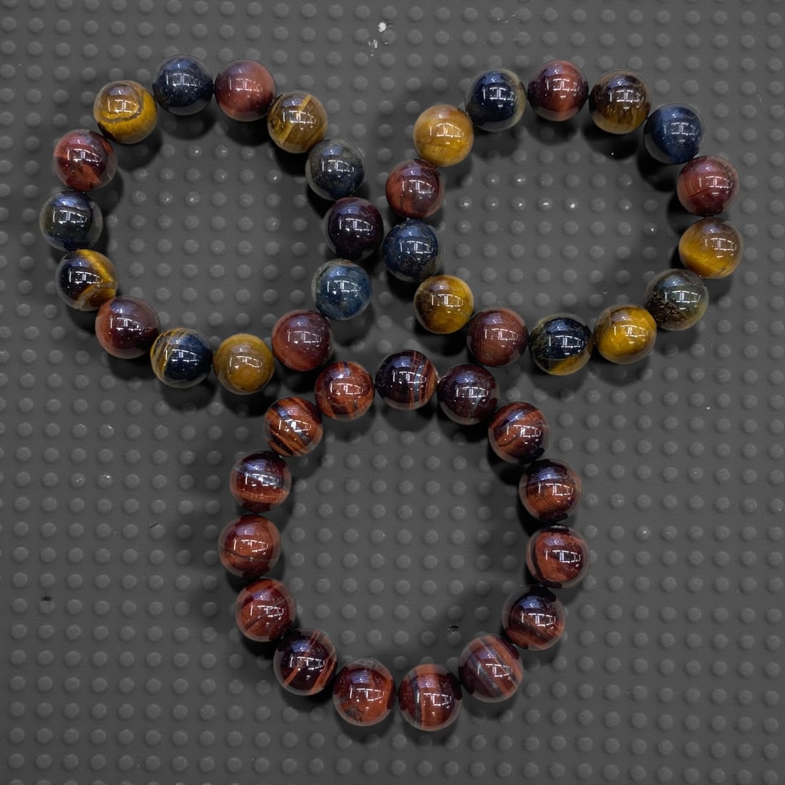 A Group of Three Tri-Color Tiger Eye Beaded Bracelets, 174g, 71AA: Each bracelet is composed of polished round tri-color tiger eye beads, displaying distinct bands of golden yellow, deep red, and iridescent blue. The beads exhibit the characteristic chatoyant effect,