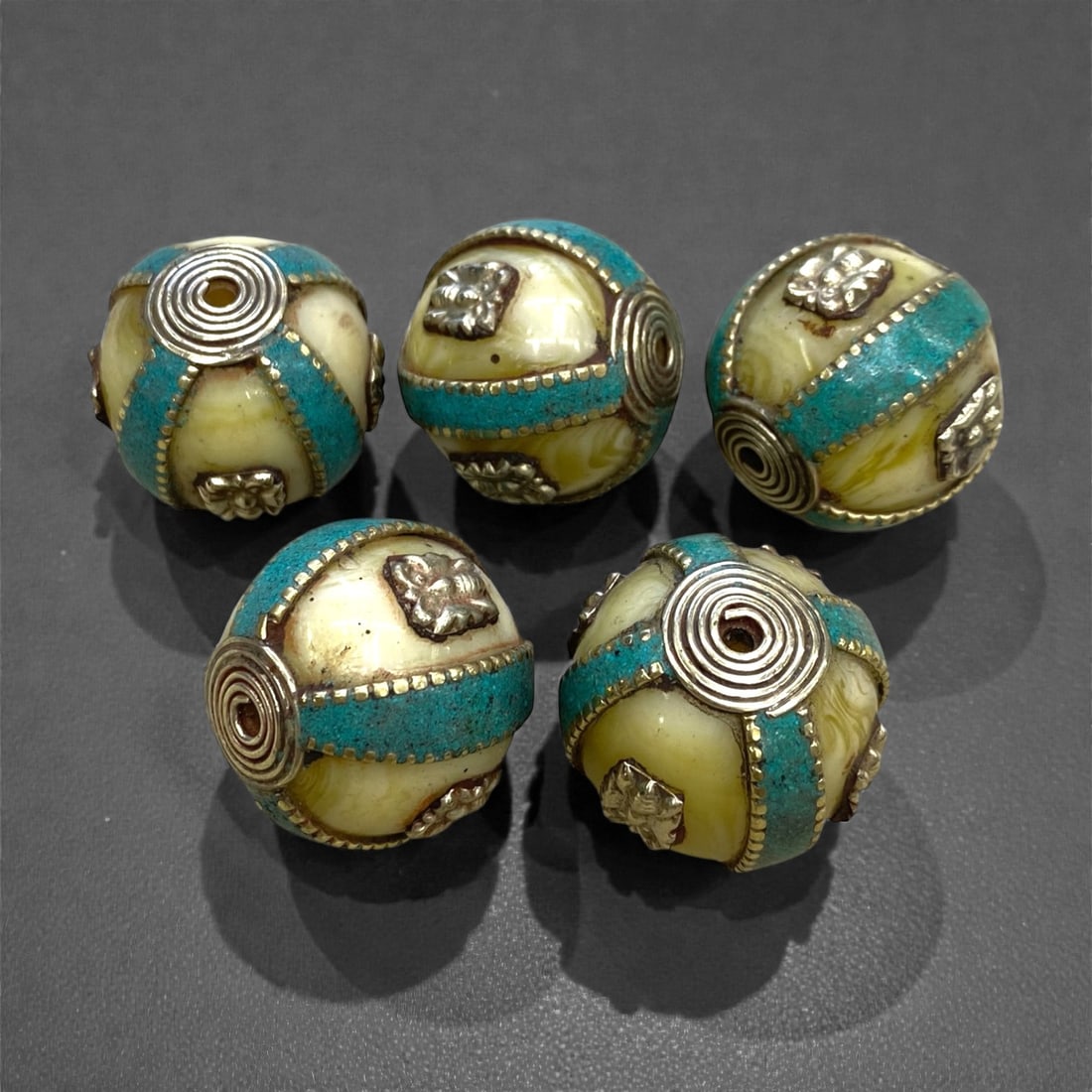 A Group of Five Tibetan Amber Resin and Silver Inlaid Beads, 20th Century, 100g, 56AA: Each bead is formed from pale yellow amber resin with subtle marbling, inlaid with bands of crushed turquoise and silver wirework. The silver elements include coiled spiral motifs and applied repousse