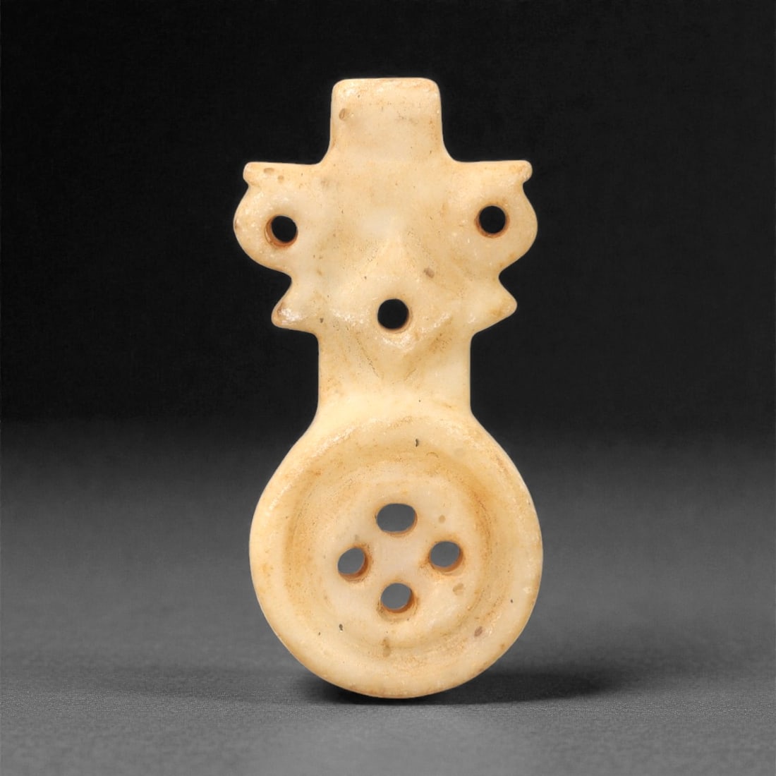 An Ancient Calcite Amulet, 2nd-1st Millennium BCE, 35FL: Carved from pale cream calcite with subtle surface patina, the object is formed as a two-part pendant. The upper section features a symmetrical, stylized design with three perforations for suspension,