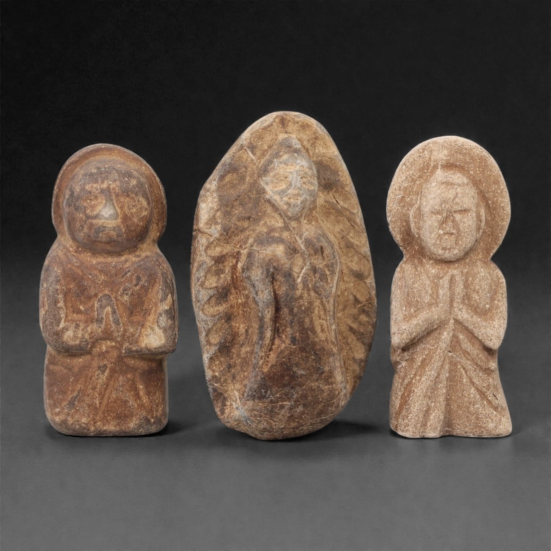 A Group of Three Japanese Stone Votive Buddha Figures, Edo Period, 50FL: Each figure is carved from light brown stone with natural surface weathering and visible fissures. The first and third figures depict standing figures with hands in anjali mudra, wearing robes and a c
