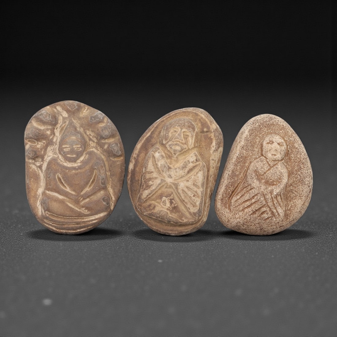 A Group of Three Japanese Stone Votive Figures, Edo Period, 55FL: Each figure is carved from light brown stone with natural surface weathering and visible patina. The first depicts a seated figure with a pointed headdress, surrounded by a radiating border. The secon