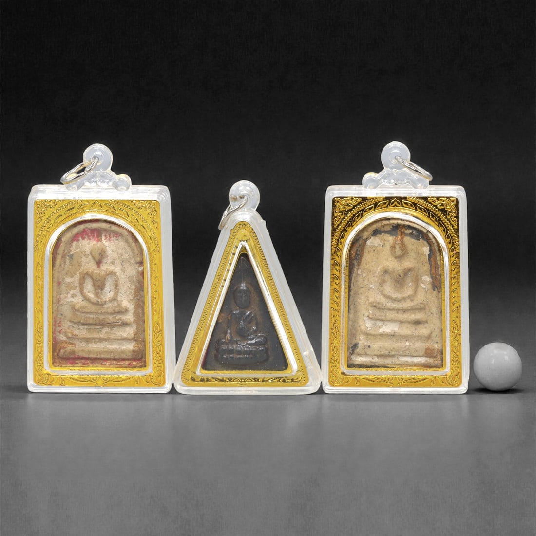 A Set of Three Thai Amulet Pendants, 20th Century, 82ZG: Encased in clear protective casings with gold-toned decorative borders, the set comprises three distinct amulet pendants, each with an integrated suspension loop for wear. The left and right pendants