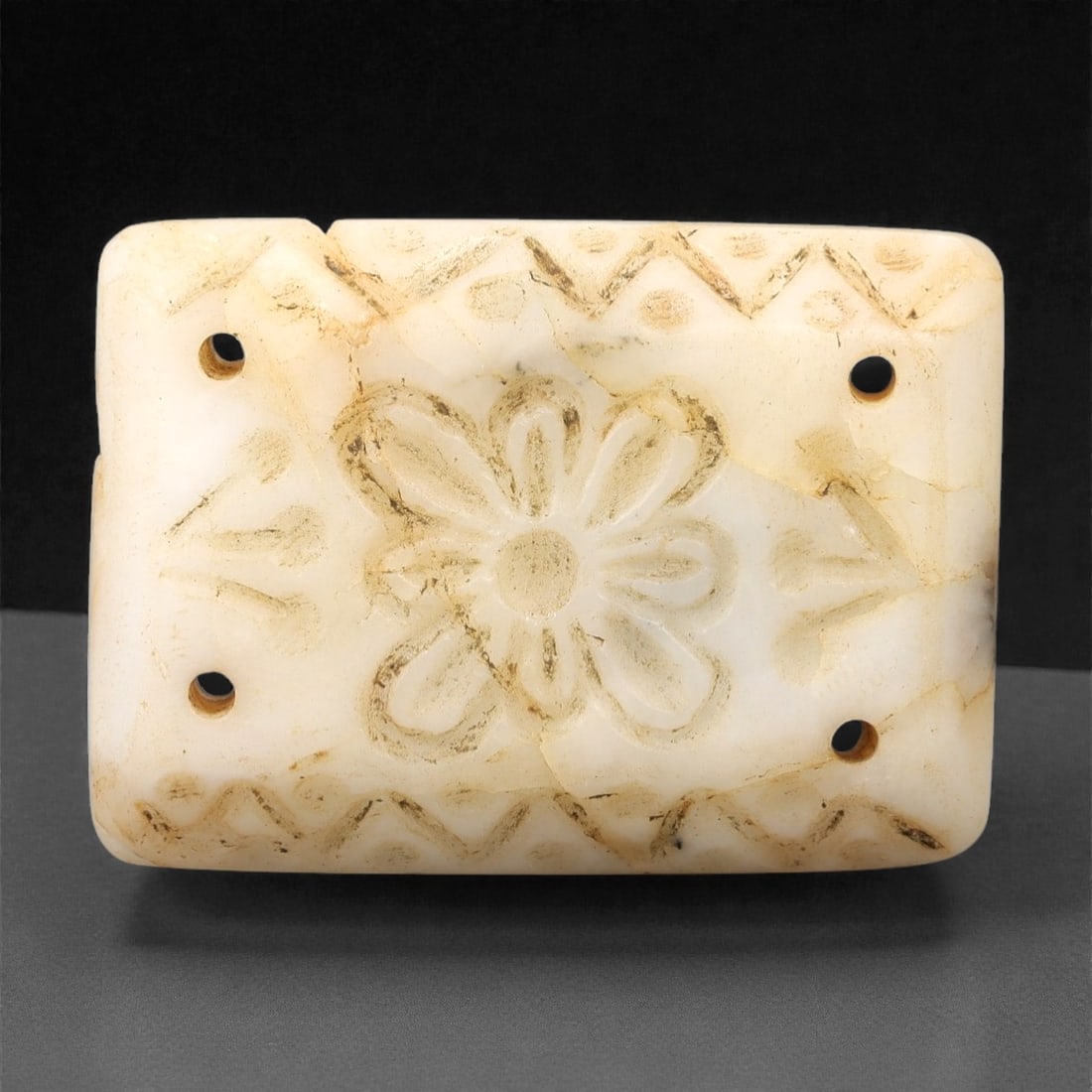 An Antique Islamic Jade Amulet, 10th-14th Century CE, 77FL: Carved from pale white jade with natural tonal variation, surface encrustation, and visible fractures, the amulet is formed as a rectangular plaque. A central floral motif is carved in relief, with ei
