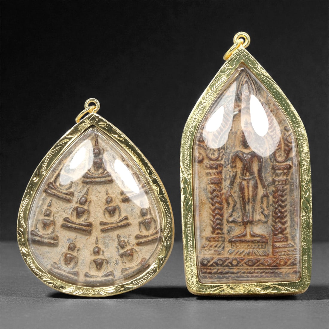 A Set of Two Thai Amulet Pendants, 20th Century, 83ZG: Encased in gold-toned metal frames with clear protective covers, the set comprises two distinct amulet pendants, each with an integrated suspension loop for wear. The left pendant is teardrop-shaped,
