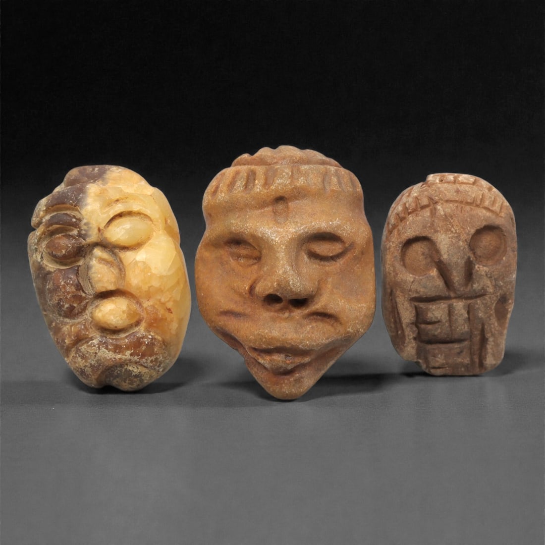 A Group of Three Medieval Stone Maskette Amulets, 10th-14th Century CE, 5ZH: Comprising three distinct maskette amulets, each carved from stone with natural color and texture variations. The first, of yellowish-brown stone with darker brown mineral inclusions and surface encru