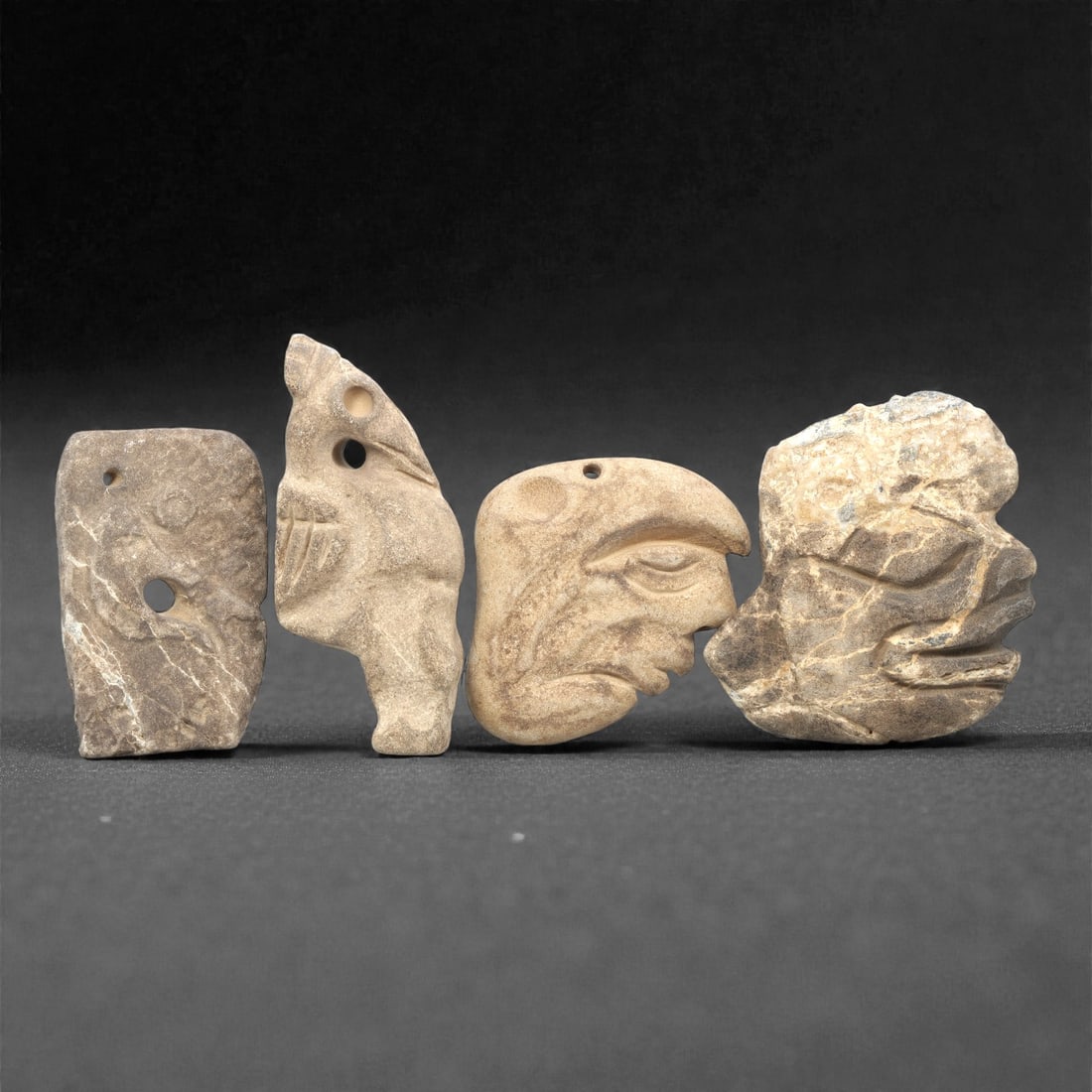 Four Pre-Columbian Stone Carvings, 10th-15th Century, 96ZG: The first carving is a rectangular pendant, formed from greyish-brown stone with visible fissures, featuring incised zoomorphic motifs and two suspension holes. The second is a zoomorphic pendant, car