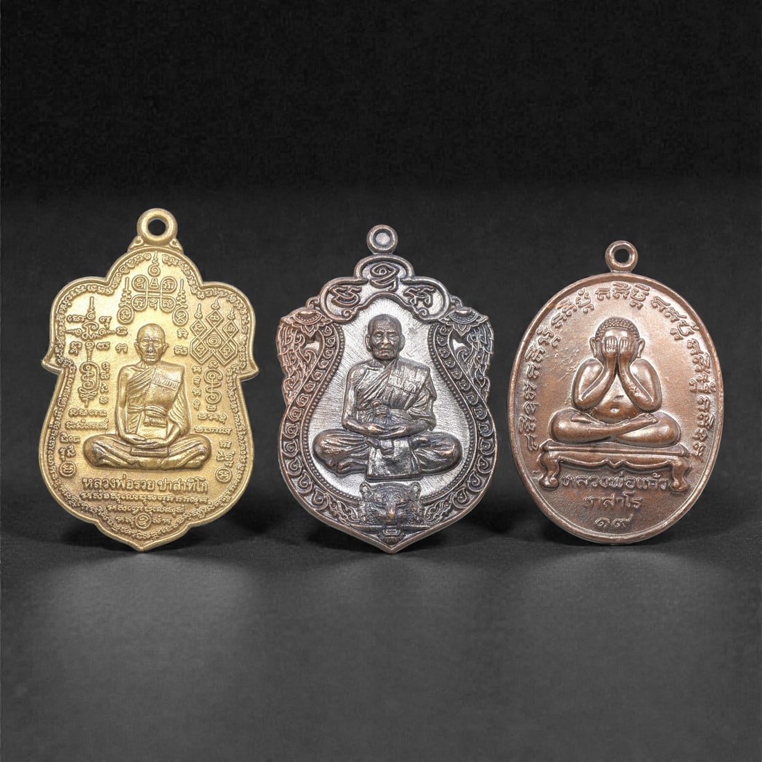 A Set of Three Thai Metal Amulet Pendants, 20th Century, 80ZG: Cast from metal with varied patination, the set comprises three distinct amulet pendants, each with an integrated suspension loop for wear. The left pendant is shield-shaped, cast in brass with a seat