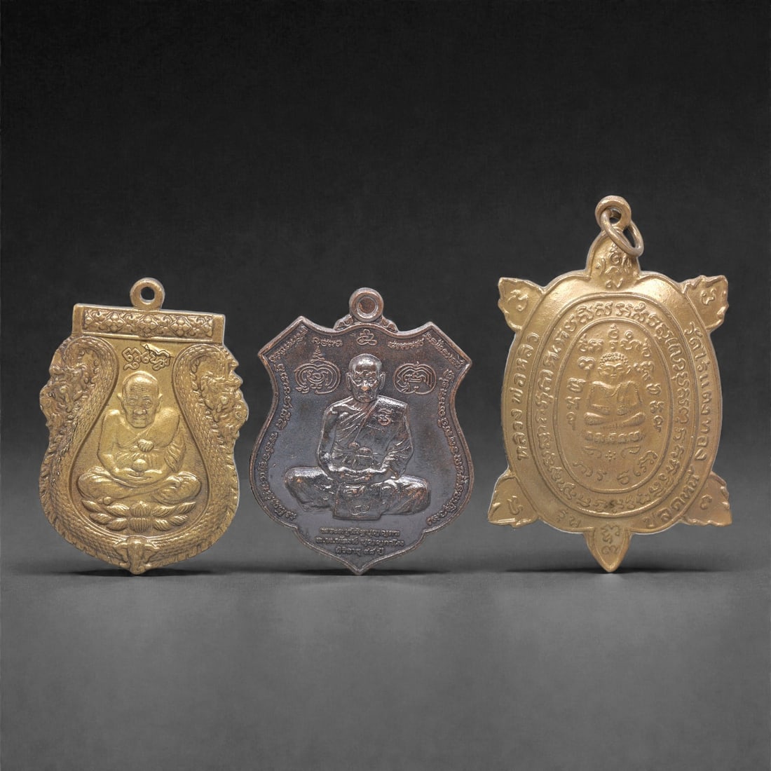 A Set of Three Thai Metal Amulet Pendants, 20th Century, 79ZG: Cast from metal with varied patination, the set comprises three distinct amulet pendants, each with an integrated suspension loop for wear. The left pendant is shield-shaped, cast in brass with a seat