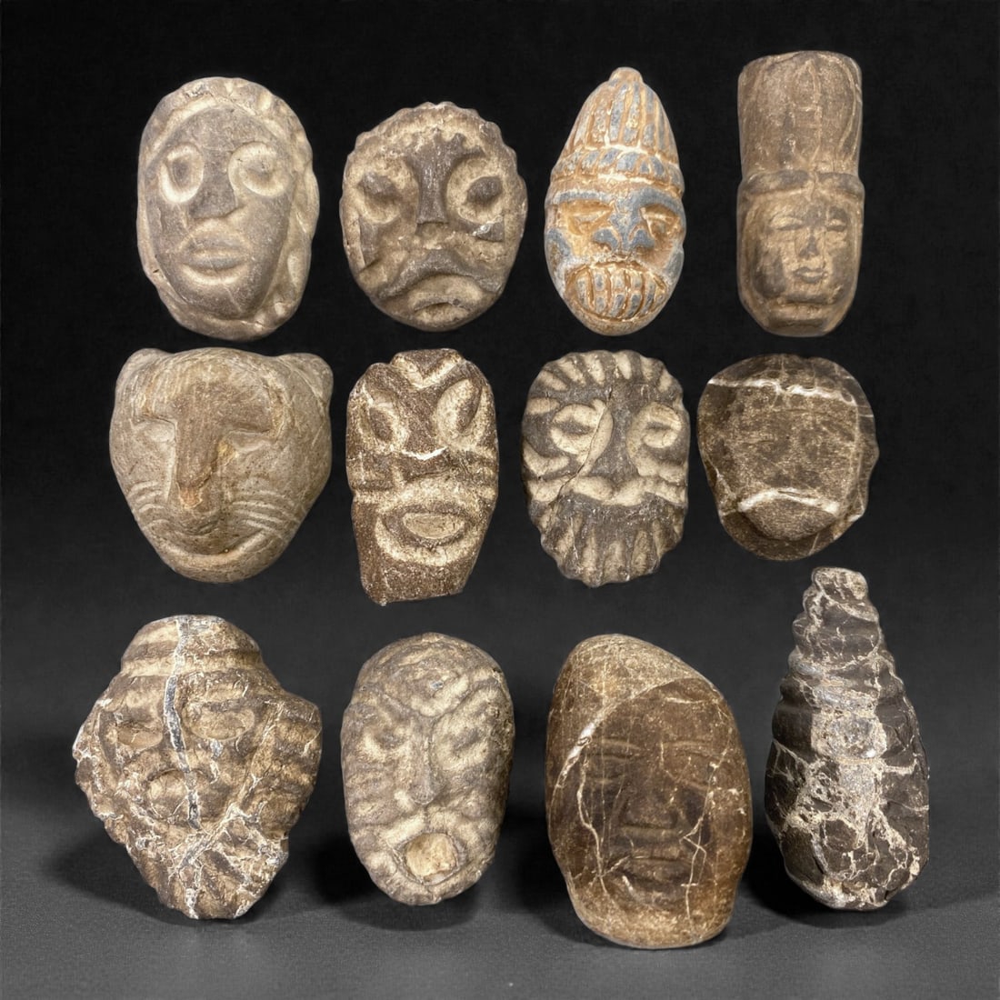 A Group of Ancient Stone Maskettes, 83Z: Carved from dense stone in tones ranging from pale beige to deep brown and charcoal, this group comprises twelve small maskettes depicting highly stylized human faces. The masks are rendered in a stro