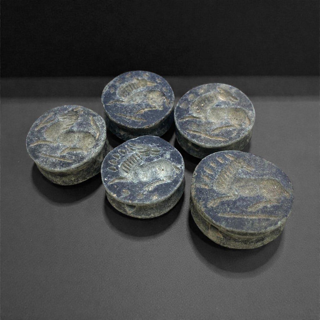 A Group of Five Near Eastern Schist Seal Beads, 3rd-2nd Millennium BCE, 85g, 51AA: Each bead is carved from fine-grained dark schist, formed as a thick disc with a central perforation for suspension. The upper face of each bead bears an incised zoomorphic motif, including recumbent