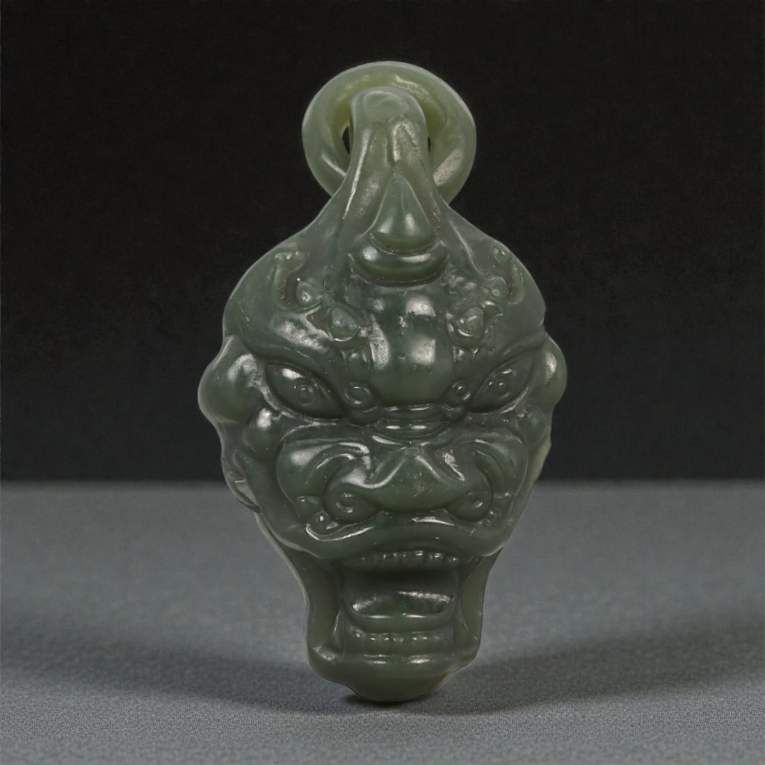 A Chinese Nephrite Jade Taotie Mask Pendant, 20th Century, 1YB: Carved from dark green nephrite jade with areas of lighter translucency and minor natural inclusions, the pendant is formed as a stylized taotie mask rendered in high relief. The face is symmetrically