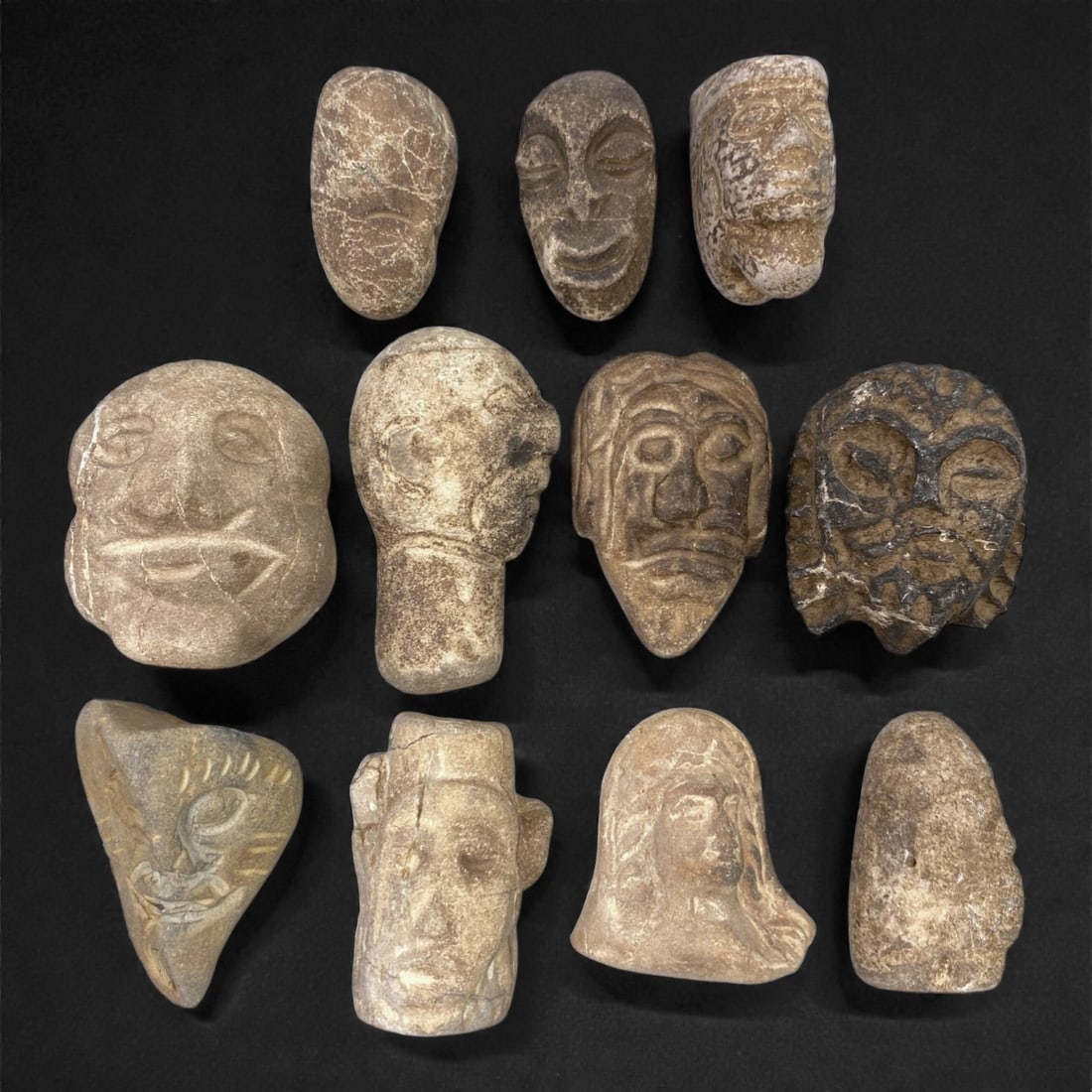 A Group of Near Eastern Stone Maskettes, Ancient Period, 70XJ: Comprising eleven small stone maskettes carved from light to dark-toned stone with natural veining and surface wear, the group consists of stylized human faces rendered in low to moderate relief, with