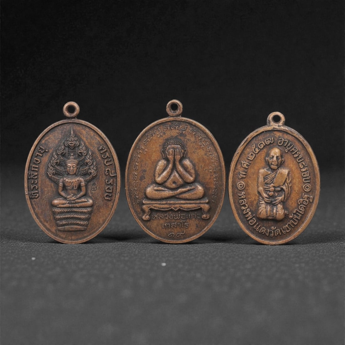 A Group of Three Thai Copper Buddhist Amulets, 20th Century, 38FL: Each amulet is cast from copper with a warm brown patina, shaped as an oval medallion with a suspension loop at the top. The first depicts a Buddha seated in meditative pose atop a coiled naga, with T