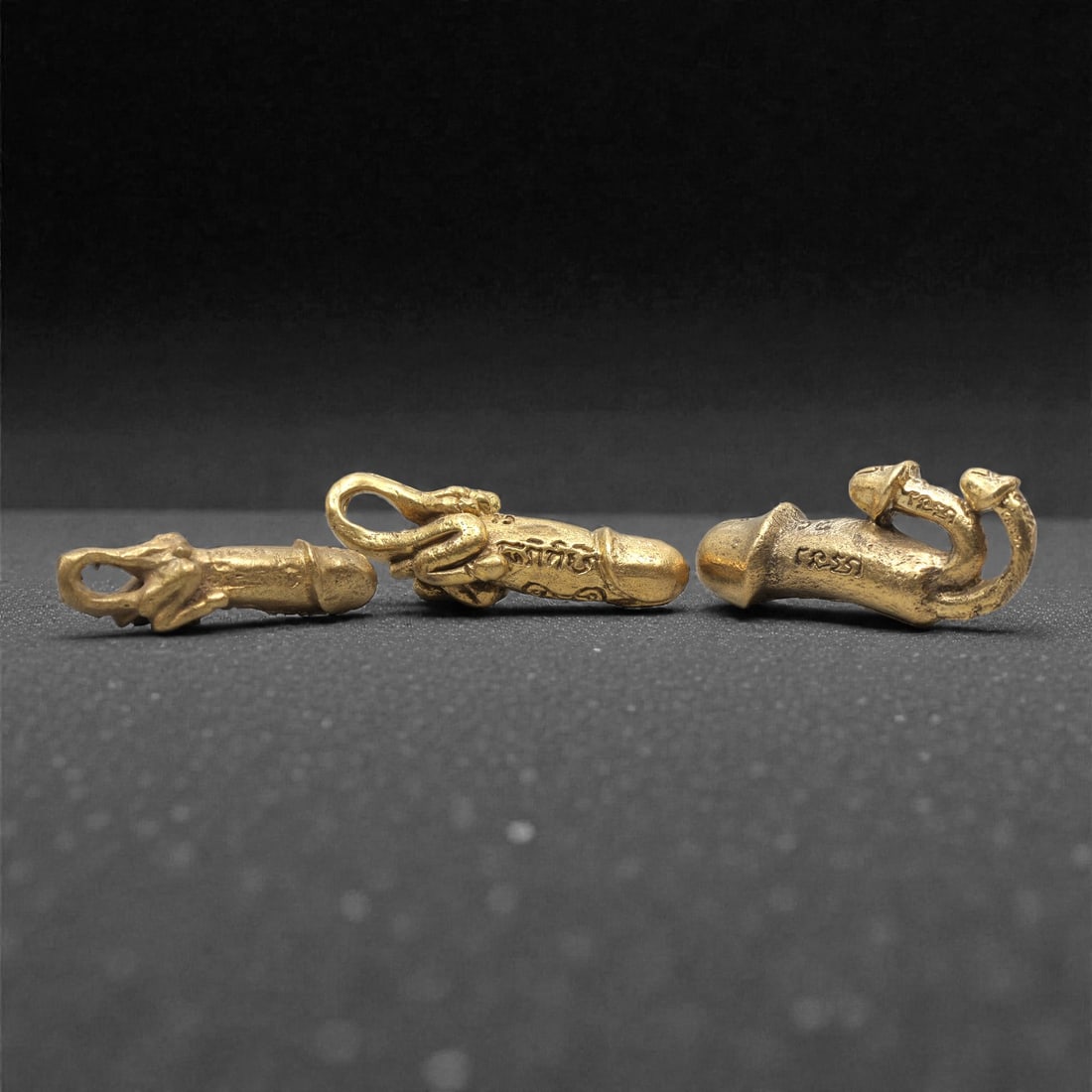 A Set of Three Thai Brass Amulet Figures, 20th Century, 61ZG: Cast from brass with a warm golden patina, the set comprises three distinct amulet figures, each with an integrated suspension loop for wear. The left figure is a stylized, elongated form with a suspe