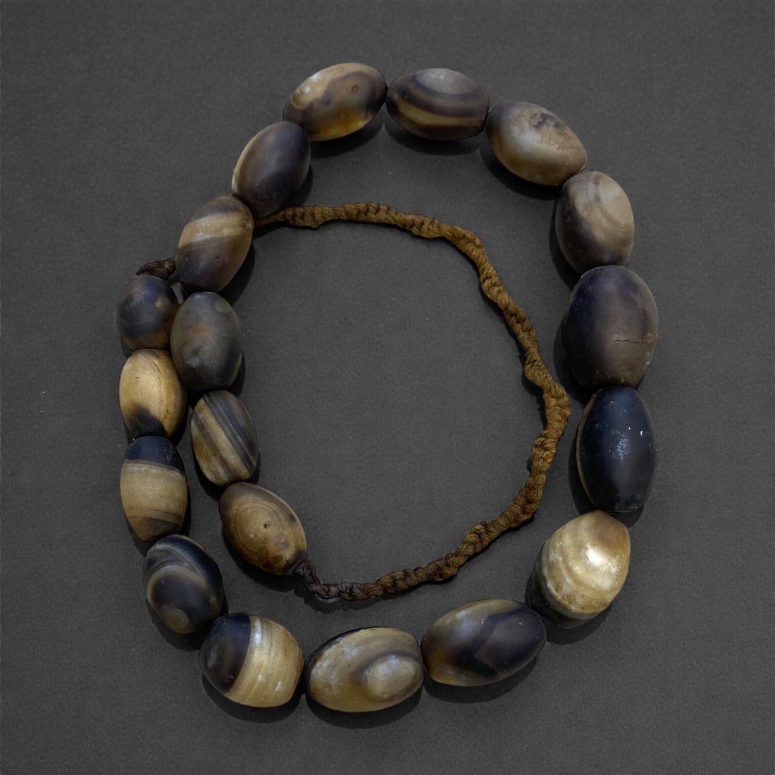A Near Eastern Banded Agate Sulaimani Bead Necklace Strand, 19th Century, 180g, 6AA: Composed of elongated oval banded agate beads with alternating layers of translucent honey-brown and opaque dark grey, strung on a modern braided fiber cord. The distinctive banding pattern is charact