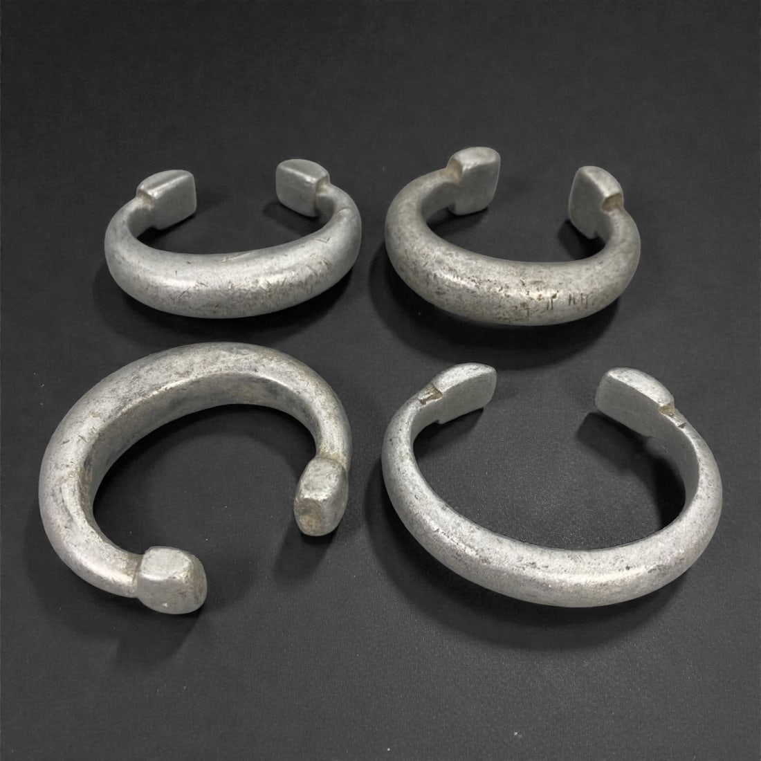 A Group of African Aluminium Bracelets, 19th-Early 20th Century, 3FL: The group comprises four cast aluminium bracelets, each formed as a thick, open hoop with squared or slightly rounded terminals and a circular to oval profile. The surfaces display extensive smoothing