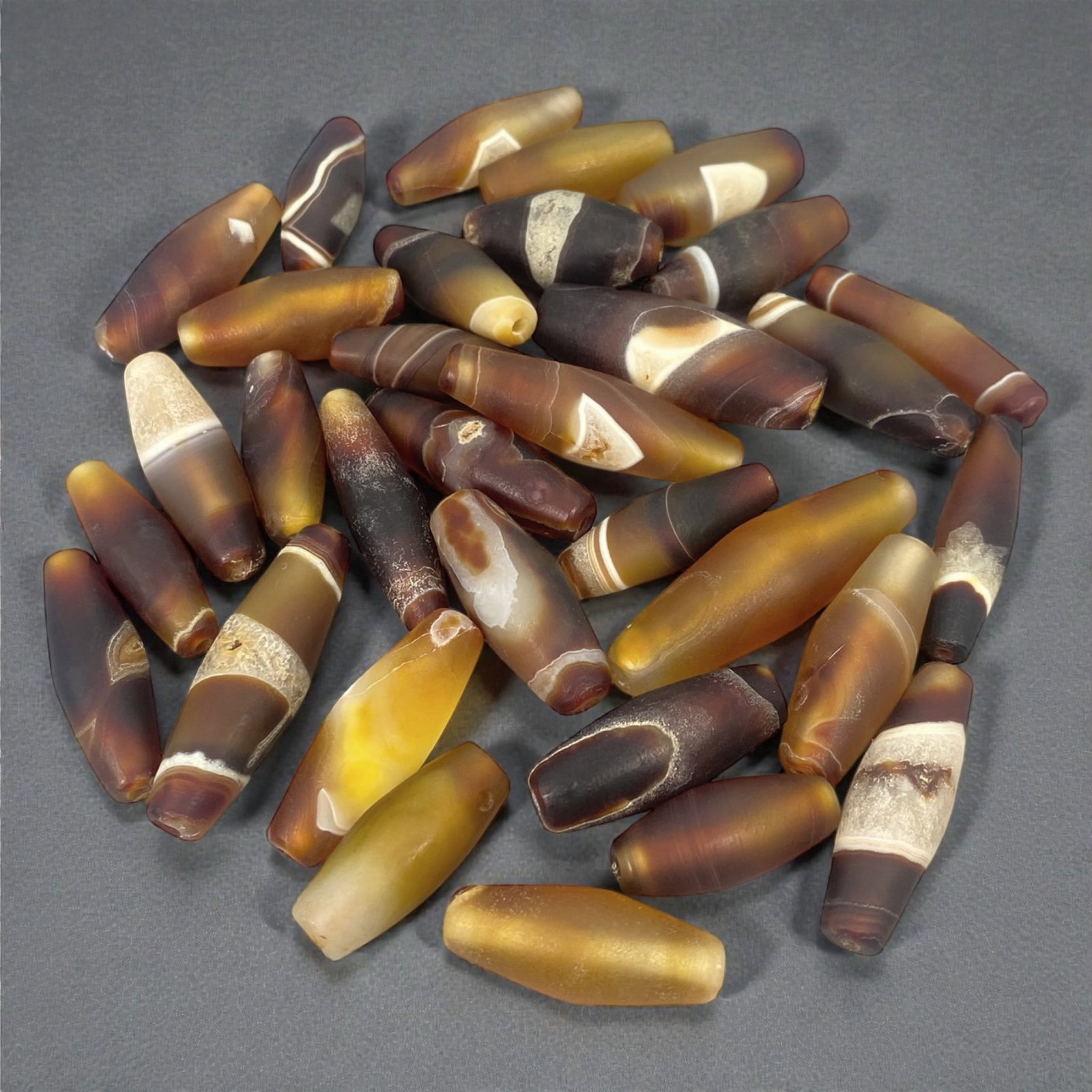 A Group of Himalayan Agate Chung Dzi Beads, 19th-20th Century CE, 11W: The group comprises thirty-two agate beads of elongated cylindrical and barrel forms, carved from translucent to semi-opaque chalcedony in tones of amber, honey, brown, cream, and dark grey. Several b