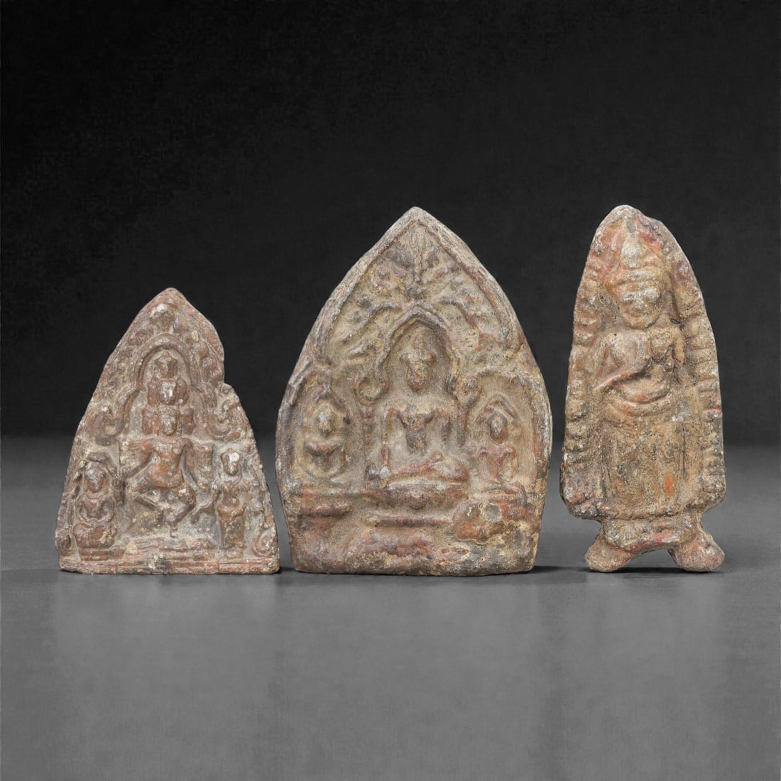 A Group of Three Thai Lead Buddhist Amulet Plaques, 20th Century, 42FL: Each amulet is formed from lead with natural surface patination, corrosion, and minor wear consistent with age and use. The leftmost plaque depicts a multi-armed deity in dynamic pose, flanked by atte
