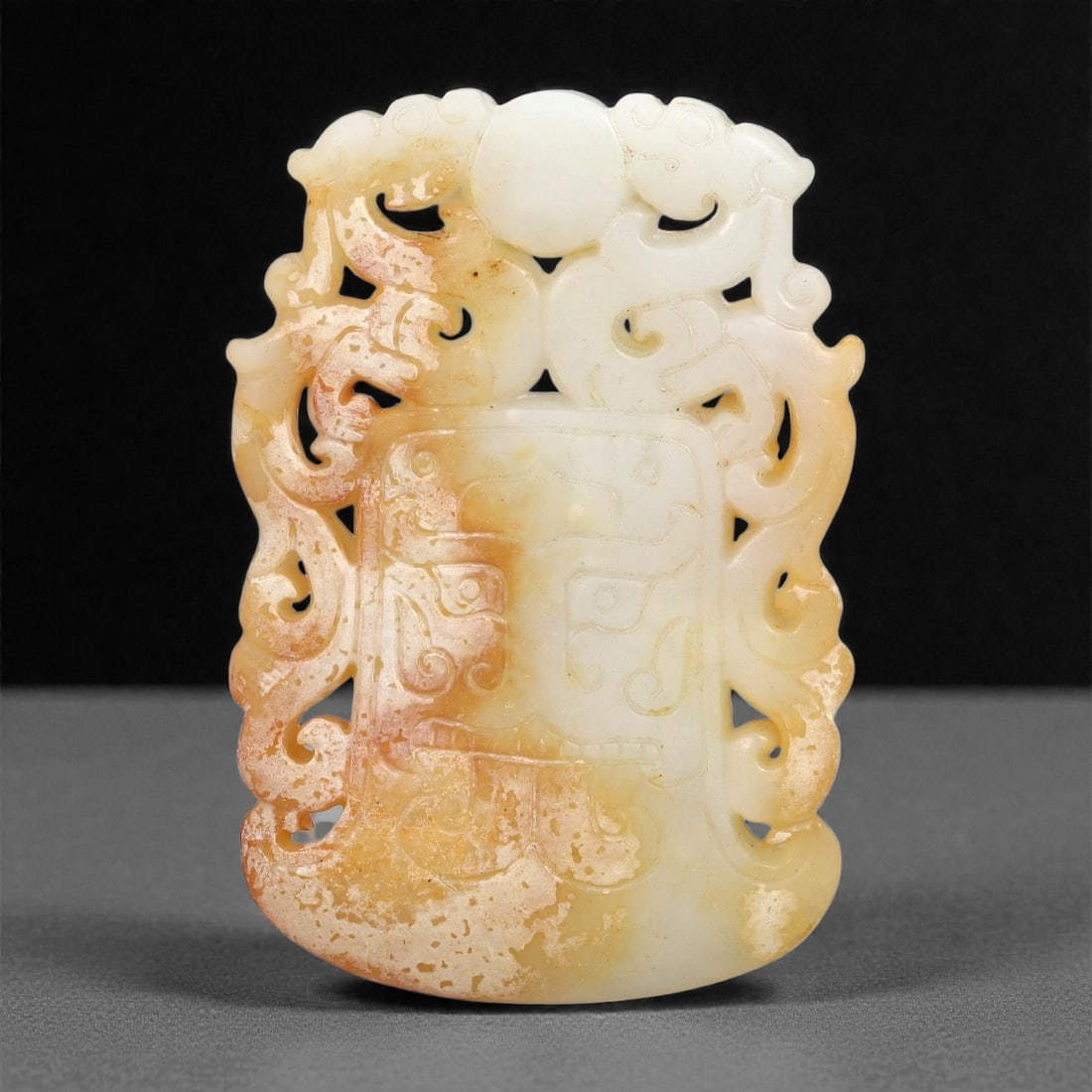 A Chinese Archaistic-Style Jade Openwork Plaque, 20th Century, 53FH: Carved from pale celadon-white jade with areas of warm russet staining and natural inclusions, the plaque is vertically oriented and executed in openwork and shallow relief. The upper section is forme
