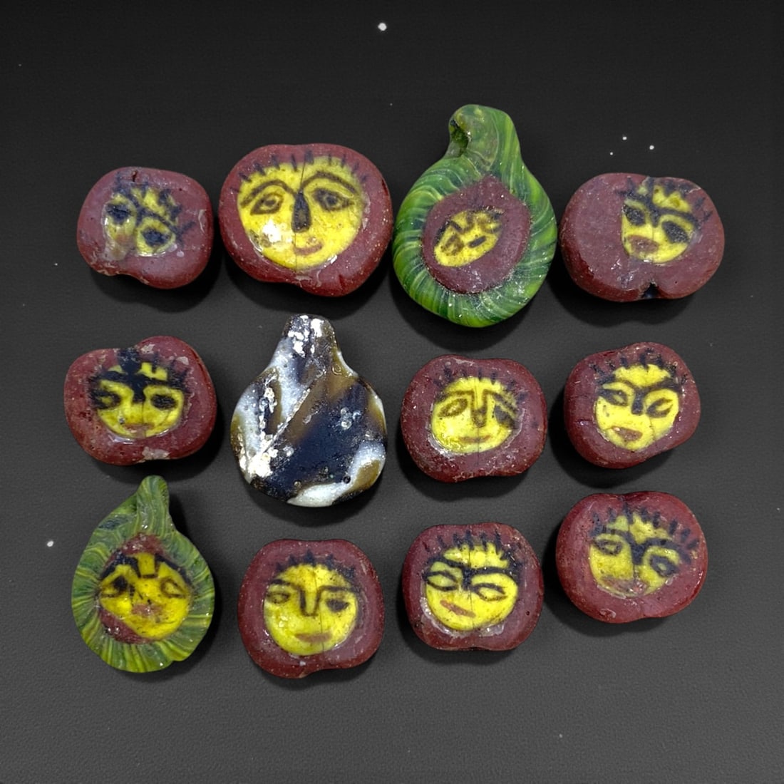 A Group of Twelve Islamic Glass Face Beads, 15g, 87AA: Comprising twelve glass face beads, primarily in a deep red glass matrix, with two examples featuring a green glass surround. Each bead is decorated with a stylized human face rendered in yellow glass