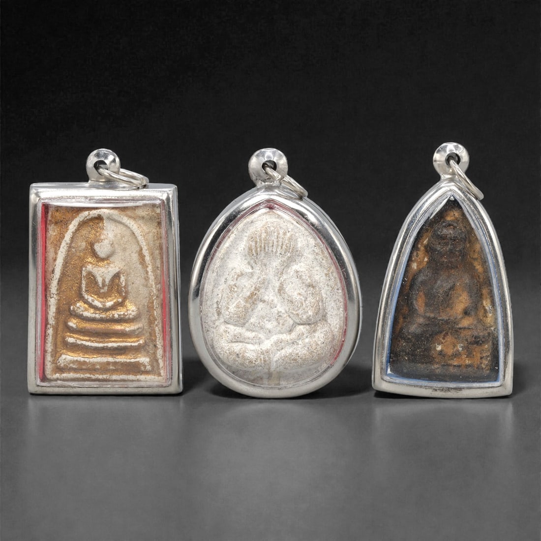 A Set of Three Thai Amulet Pendants, 20th Century, 81ZG: Encased in silver-toned metal frames, the set comprises three distinct amulet pendants, each with an integrated suspension loop for wear. The left pendant is rectangular, with a beige-toned relief Bud