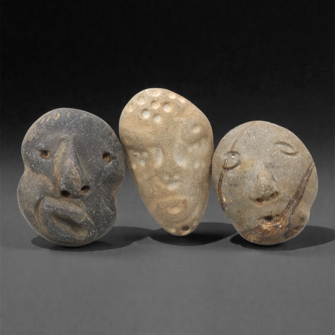 A Group of Three Medieval Stone Maskette Amulets, 10th-14th Century CE, 8ZH: Comprising three distinct maskette amulets, each carved from stone with natural color and texture variations. The first, of dark grey stone with a granular surface, is rounded with clearly defined eye