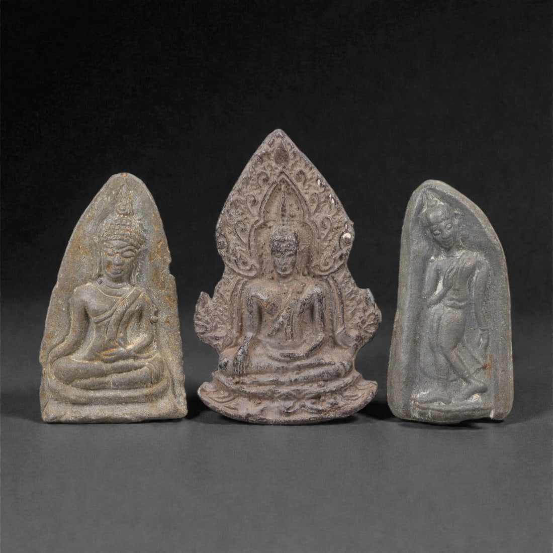 A Group of Three Thai Metal Buddhist Amulet Plaques, 20th Century, 41FL: Each amulet is formed from a dark metal alloy with natural surface patination, corrosion, and minor wear consistent with age and use. The left plaque depicts a seated Buddha in bhumisparsha mudra with