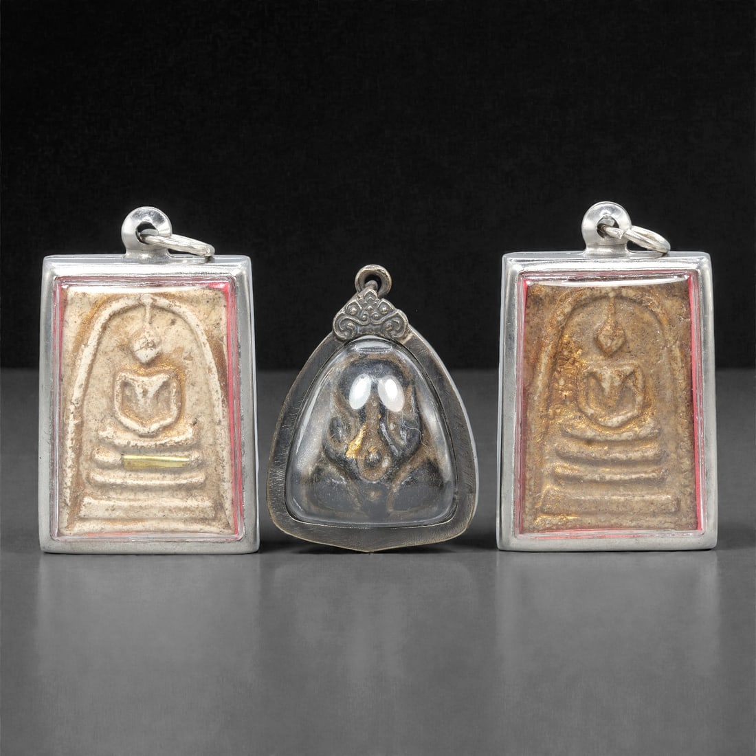 A Set of Three Thai Amulet Pendants, 20th Century, 84ZG: Encased in metal frames with clear protective covers, the set comprises three distinct amulet pendants, each with an integrated suspension loop for wear. The left and right pendants are rectangular, e