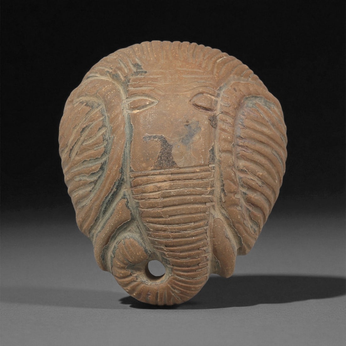 An Indo-Pacific Stone Elephant-Head Plaque, 19th-20th Century, 46ZA: Carved from a light brown stone with darker surface inclusions, the plaque is shaped as a stylized elephant head with prominent ears defined by deeply cut curving grooves. The eyes are rendered as nar