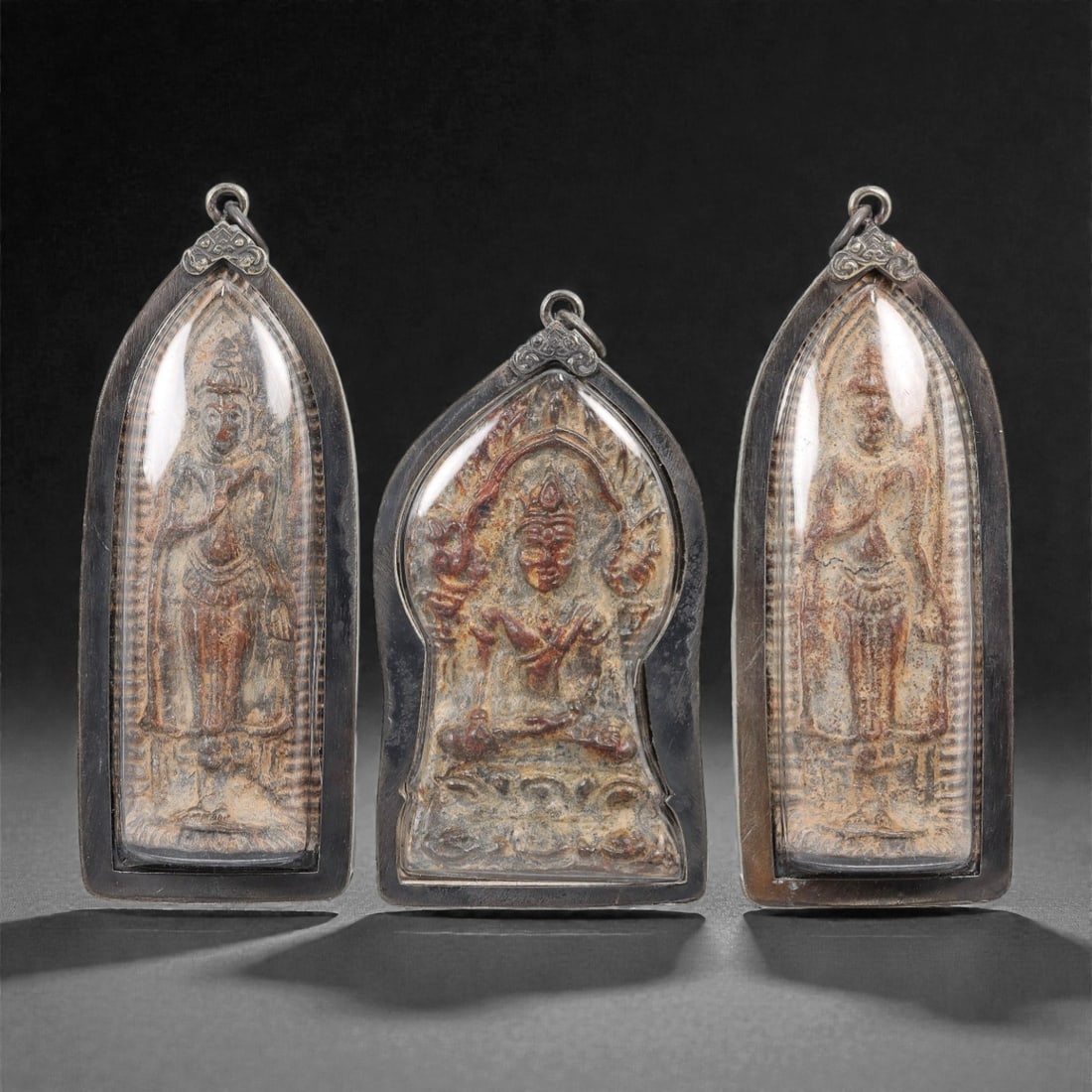 A Group of Three Thai Metal Buddhist Amulets, 20th Century, 23FL: Each amulet is formed from a metal alloy with natural surface patination and minor wear, enclosed in a dark metal case with a suspension loop for wear. The left and right amulets depict a standing dei