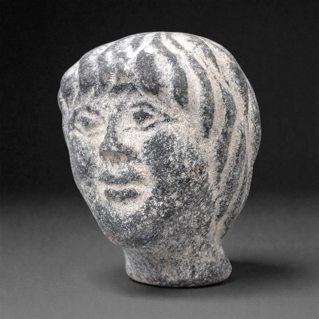 An Ancient Greco-Roman Stone Head Bust, 1st-3rd Century CE, 72FL: Carved from dark grey stone with extensive light grey and white surface encrustation, the bust depicts a human head in a naturalistic style. The hair is rendered with deep, incised wavy lines, framing