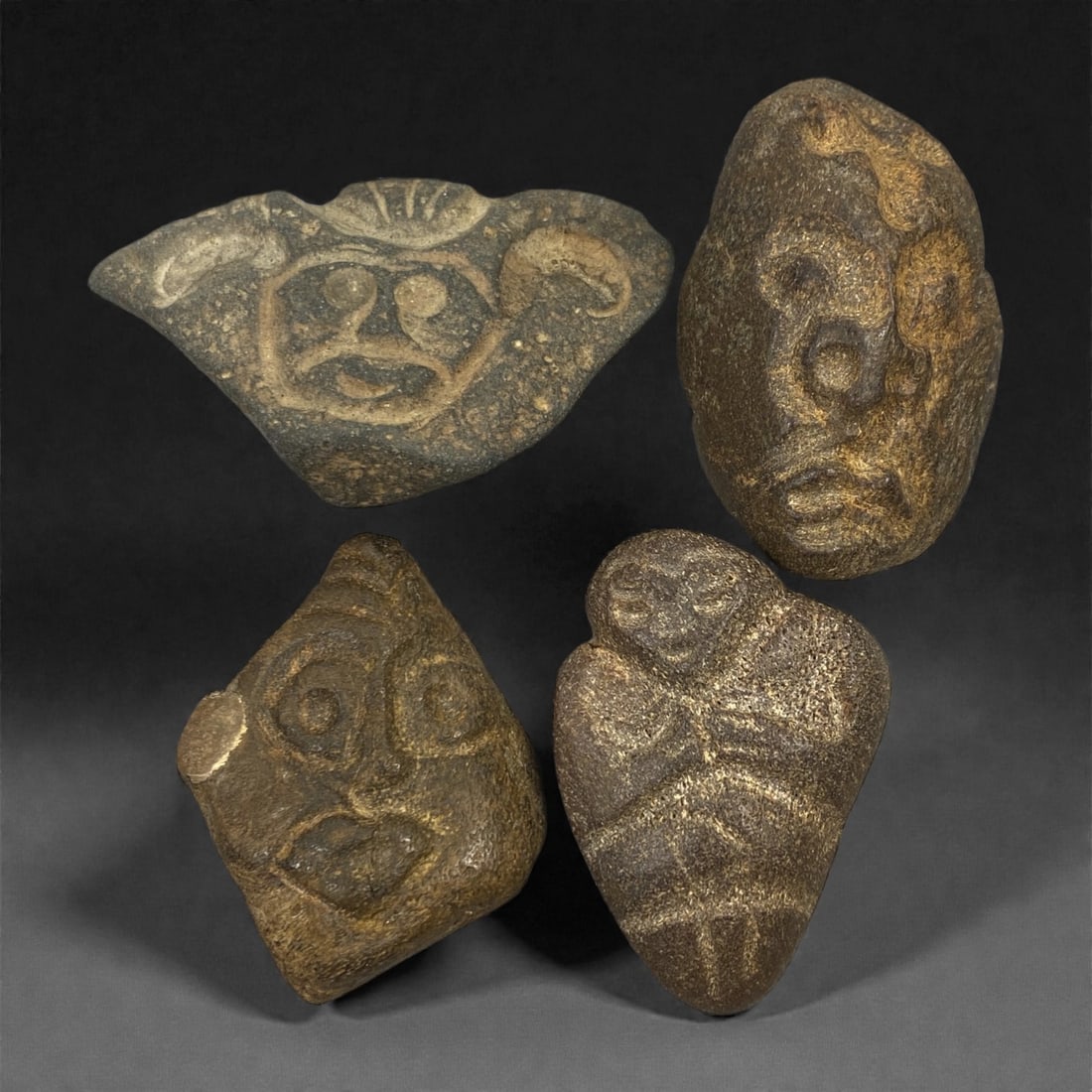 A Group of Four Ancient Stone Busts, 1st Millennium BCE, 15FN: Carved from stone with natural patination, mineral inclusions, and surface wear, the four busts depict humanoid and zoomorphic forms in low to medium relief. The top-left example is fan-shaped, featur