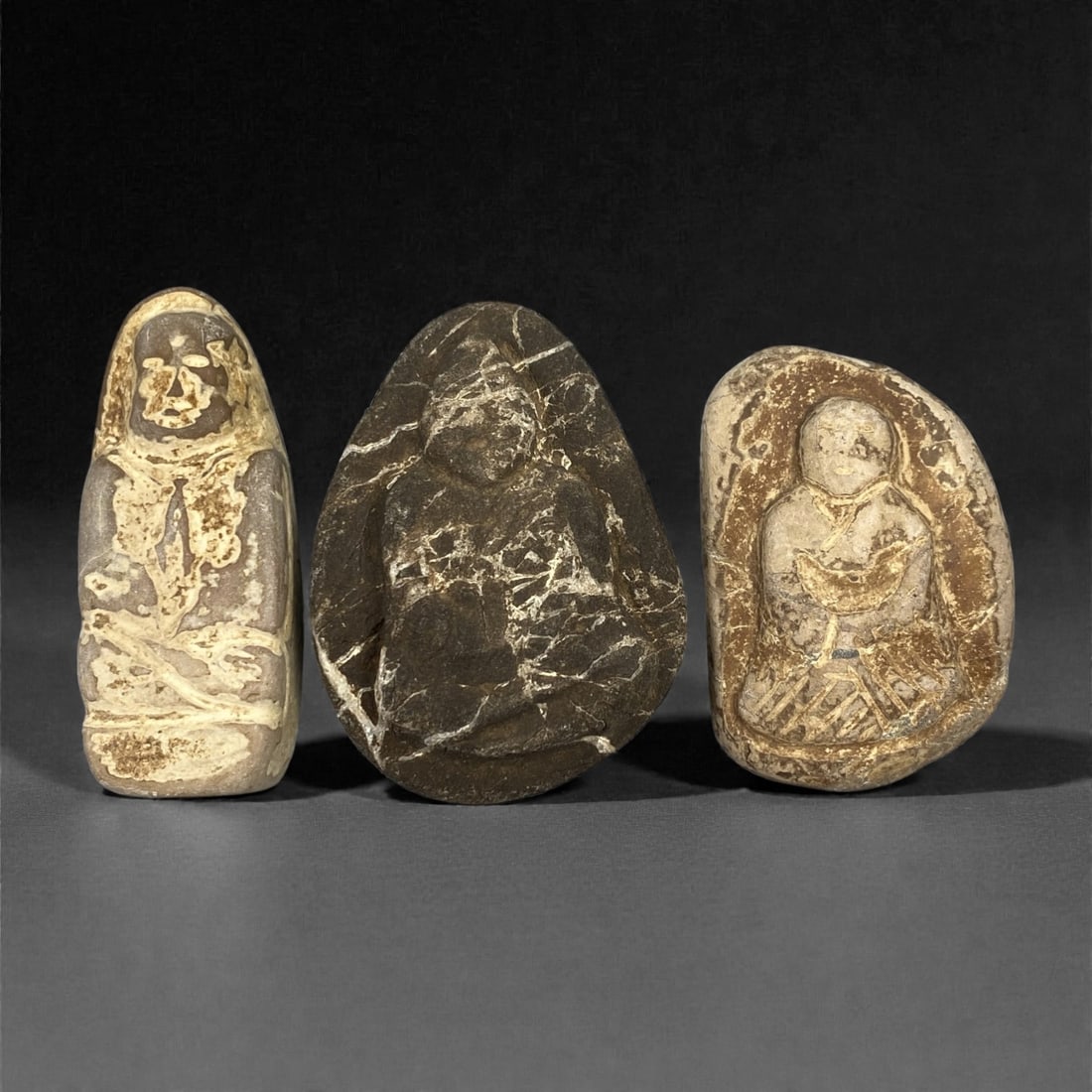 A Group of Three Edo Period Buddhist Stone Carvings, 17th-19th Century, 31FN: Carved from stone with natural tonal and textural variation, the three objects depict seated Buddhist figures in a meditative posture. The leftmost figure is vertically oriented, with a simplified fac