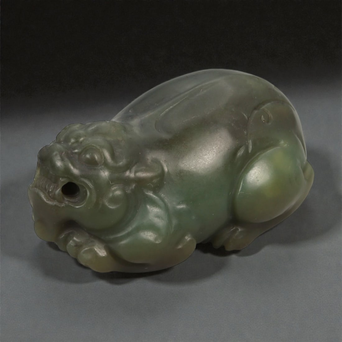 A Chinese Jade Mythical Beast Figure, 20th Century, 89FB: Carved from dark green jade with areas of lighter translucency and subtle tonal variation, the figure is worked in the round as a recumbent mythical beast shown resting on its haunches. The compact bo