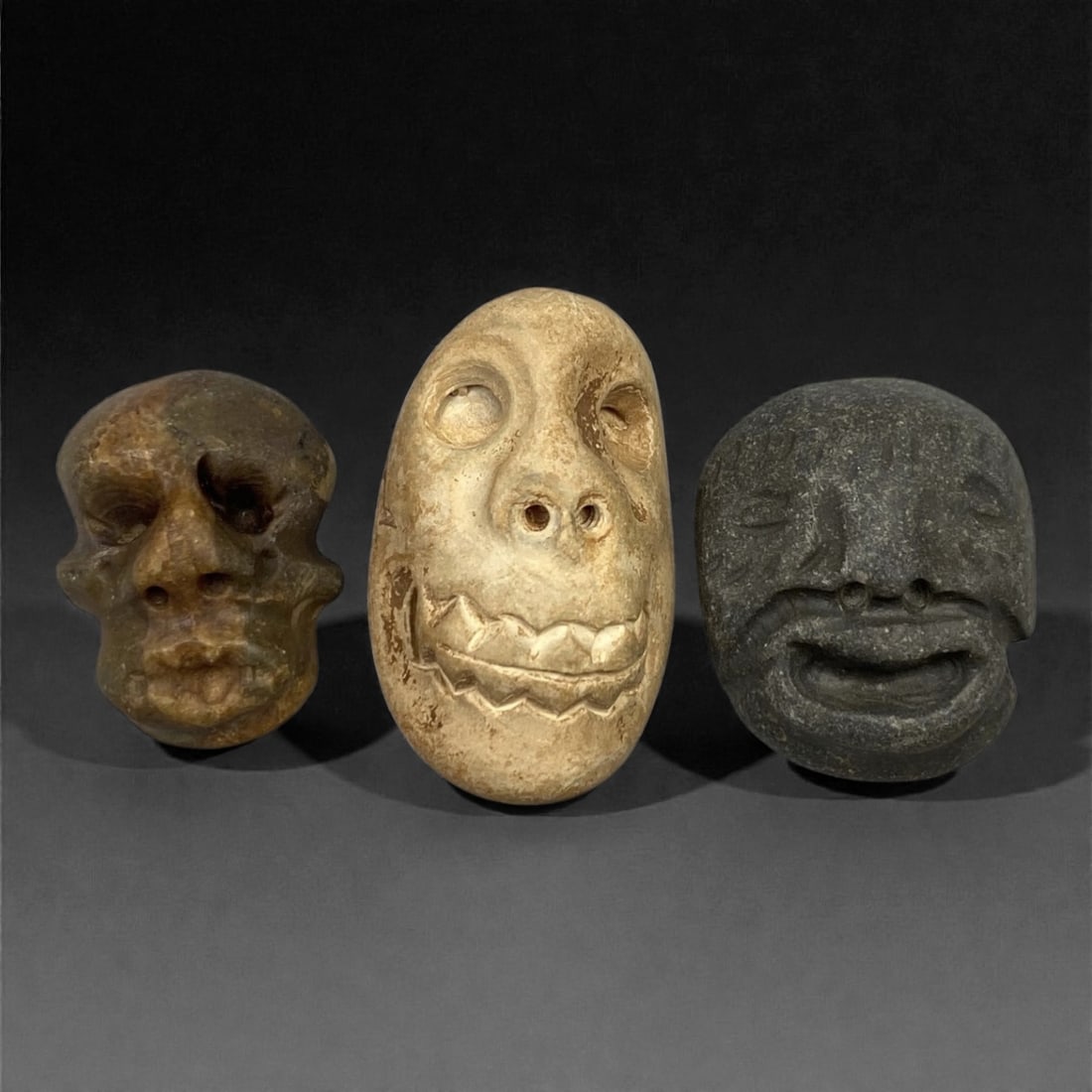 A Group of Three Medieval Stone Maskette Amulets, 10th-14th Century, 9FM: Carved from stone with varied natural patination, the set comprises three miniature maskette amulets: one in brownish stone with a skull-like form, one in light beige stone with prominent teeth and ho