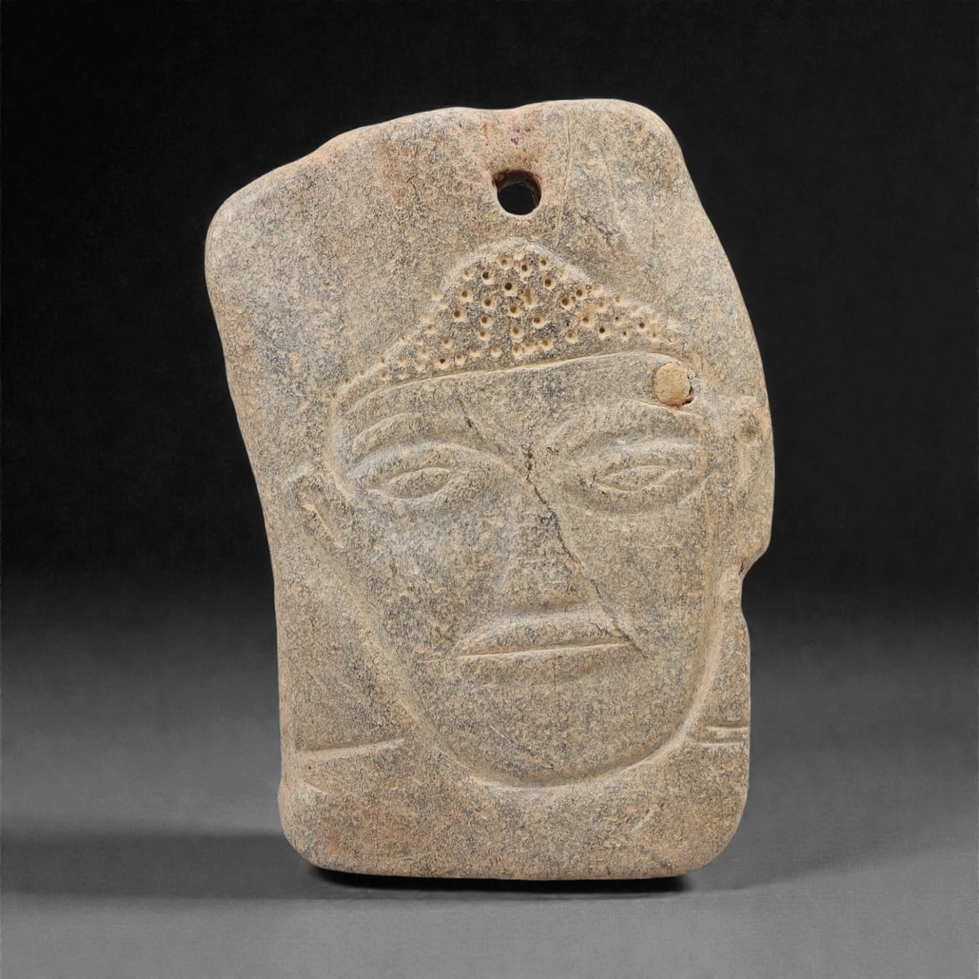 A Sino-Siberian Stone Relief Plaque, 1st Millennium BCE, 88FM: Carved from grey-brown stone with natural mottling and surface weathering, the rectangular plaque depicts a human face in shallow relief. The head is adorned with a headdress featuring rows of small p