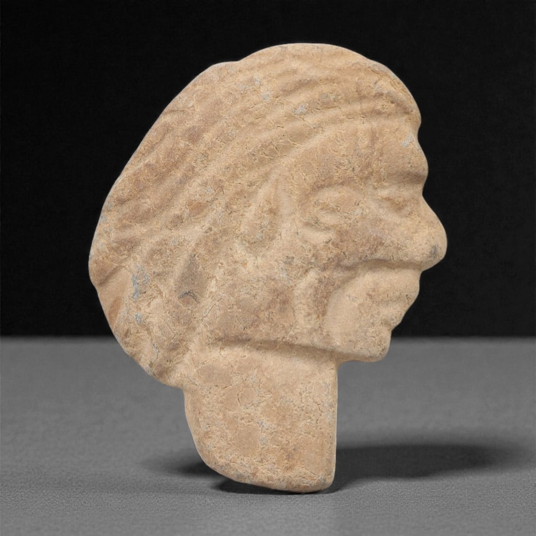 A Near Eastern Stone Head Fragment, 2nd-1st Millennium BCE, 46FM: Carved from light beige stone with a porous, slightly weathered surface and minor surface abrasions, the object is a fragmentary head in profile, featuring stylized hair rendered with incised linear d