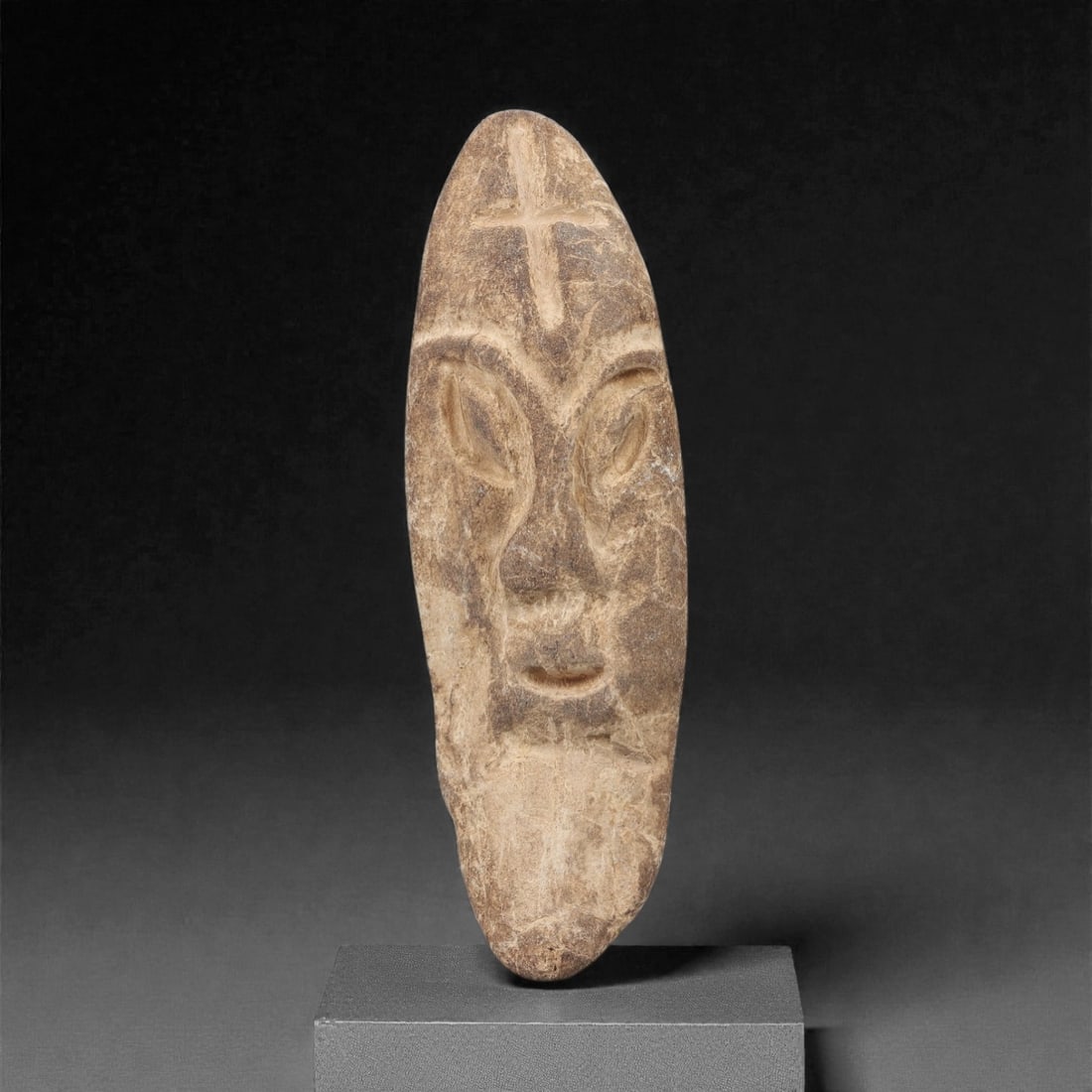 A Near Eastern Stone Head Effigy, 3rd-2nd Millennium BCE, 86FL: Carved from light brown stone with natural surface patination and weathering, the object is formed as an elongated, teardrop-shaped head effigy. The face is rendered in low relief with stylized eyes,