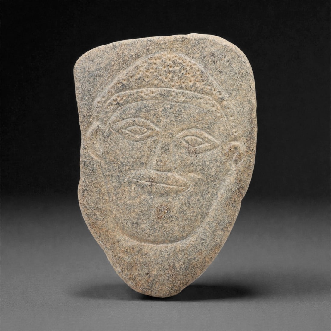 A Sino-Siberian Stone Relief Plaque, 1st Millennium BCE, 89FM: Carved from grey-brown stone with natural mottling and surface weathering, the irregularly shaped plaque depicts a human face in shallow relief. The head is adorned with a headdress featuring incised
