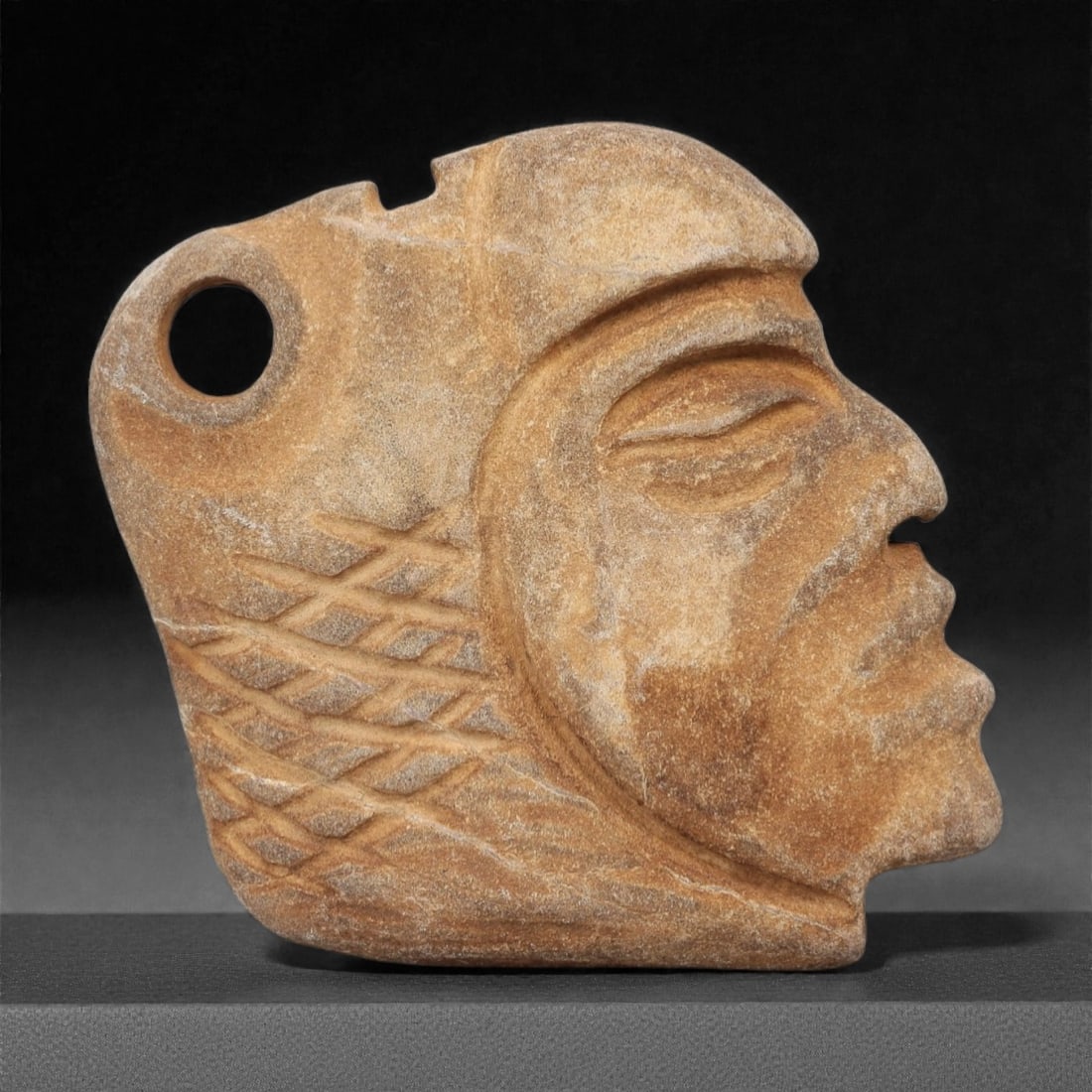 A Pre-Columbian Stone Head Pendant, 10th-15th Century, 39FM: Carved from light brown stone with a slightly weathered surface and natural tonal variation, the object is formed as a head pendant in profile, featuring a prominent brow, recessed eye socket, defined