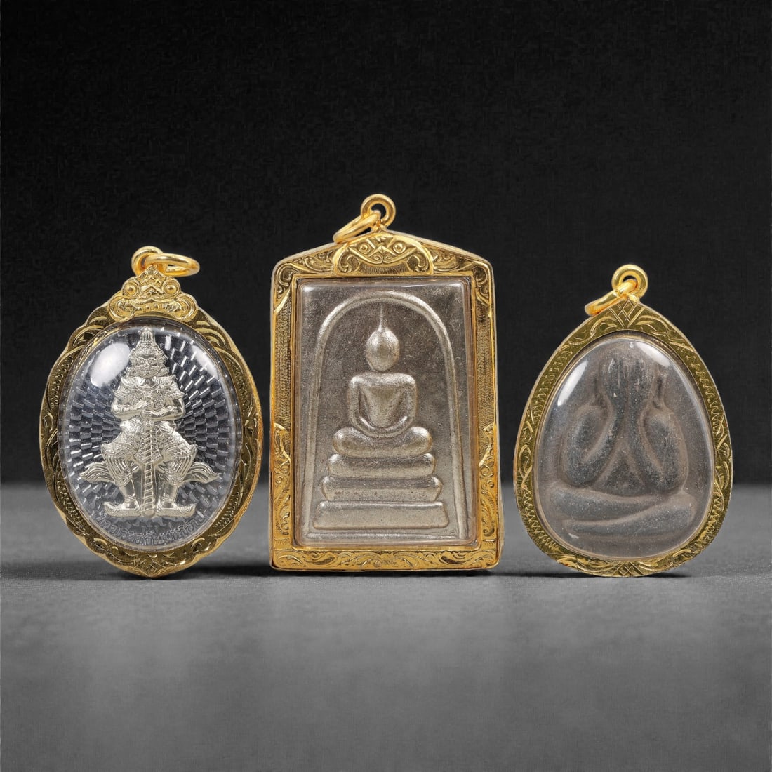A Group of Three Thai Buddhist Amulets, 20th Century, 93FM: Comprising three Thai Buddhist amulets, each encased in ornate metal frames with visible surface patina and wear. The leftmost amulet is an oval tablet, featuring a silver relief of the guardian deity