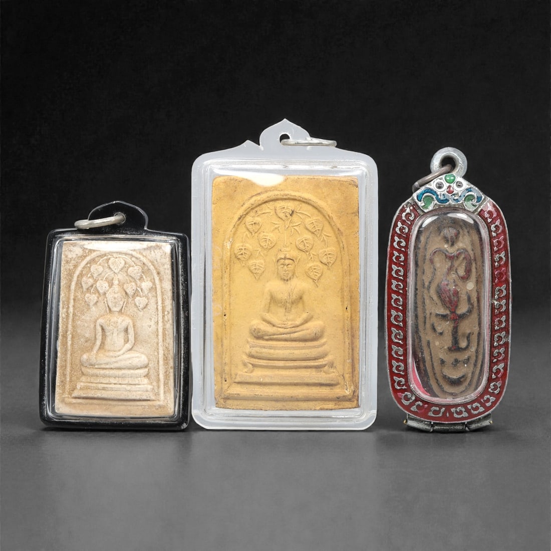 A Group of Three Thai Terracotta Amulets, 20th Century, 81XJ: Each amulet is formed from terracotta, enclosed in a case with a suspension loop for wear. The left and central amulets depict a seated Buddha in bhumisparsha mudra, with a tiered base below and a bod