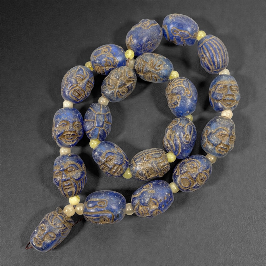 A Necklace of Blue Face Beads, 20th Century, 40FJ: The necklace is composed of carved oval beads rendered in a deep cobalt-blue material, identified as amber resin, each bead individually worked with stylized human faces, masks, and abstract linear mo