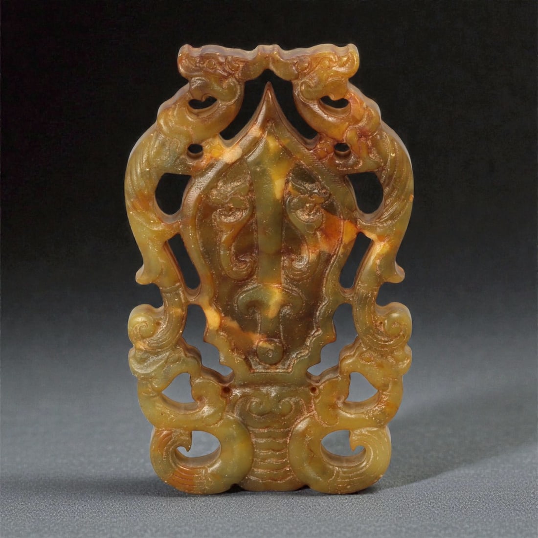 A Chinese Hardstone Openwork Plaque with Archaistic Mask Motif, 20th Century, 8YB: Carved from a mottled hardstone of translucent green, amber, and russet tones, the plaque is worked in openwork and low relief as a vertically oriented composition. The central field is dominated by a
