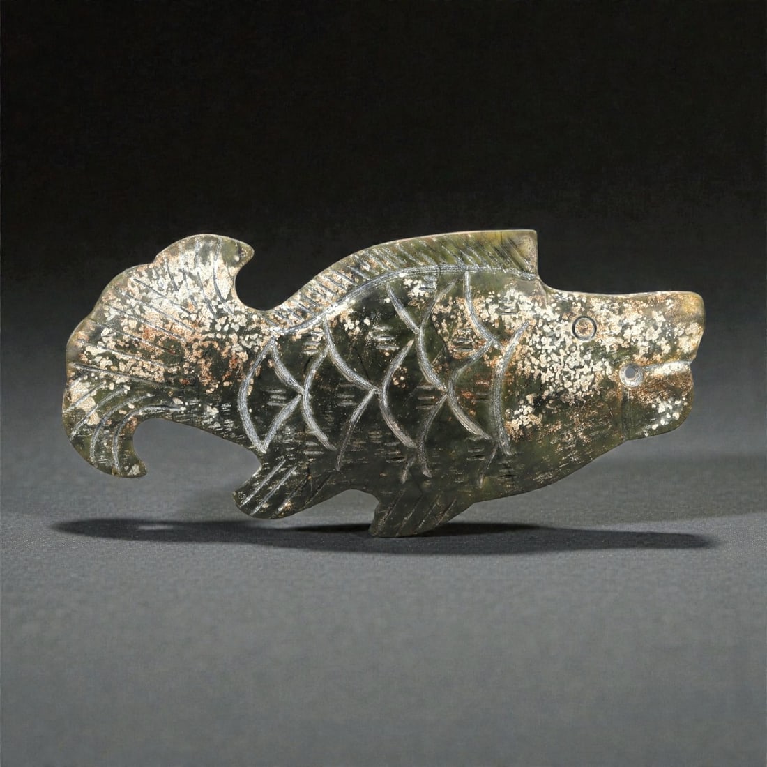 A Chinese Nephrite Jade Fish Ornament, 20th Century, 26FB: Carved from dark green Hetian nephrite jade with extensive natural inclusions and mottled surface mineralization, the ornament is worked in the form of a fish shown in profile. The body is carved in l