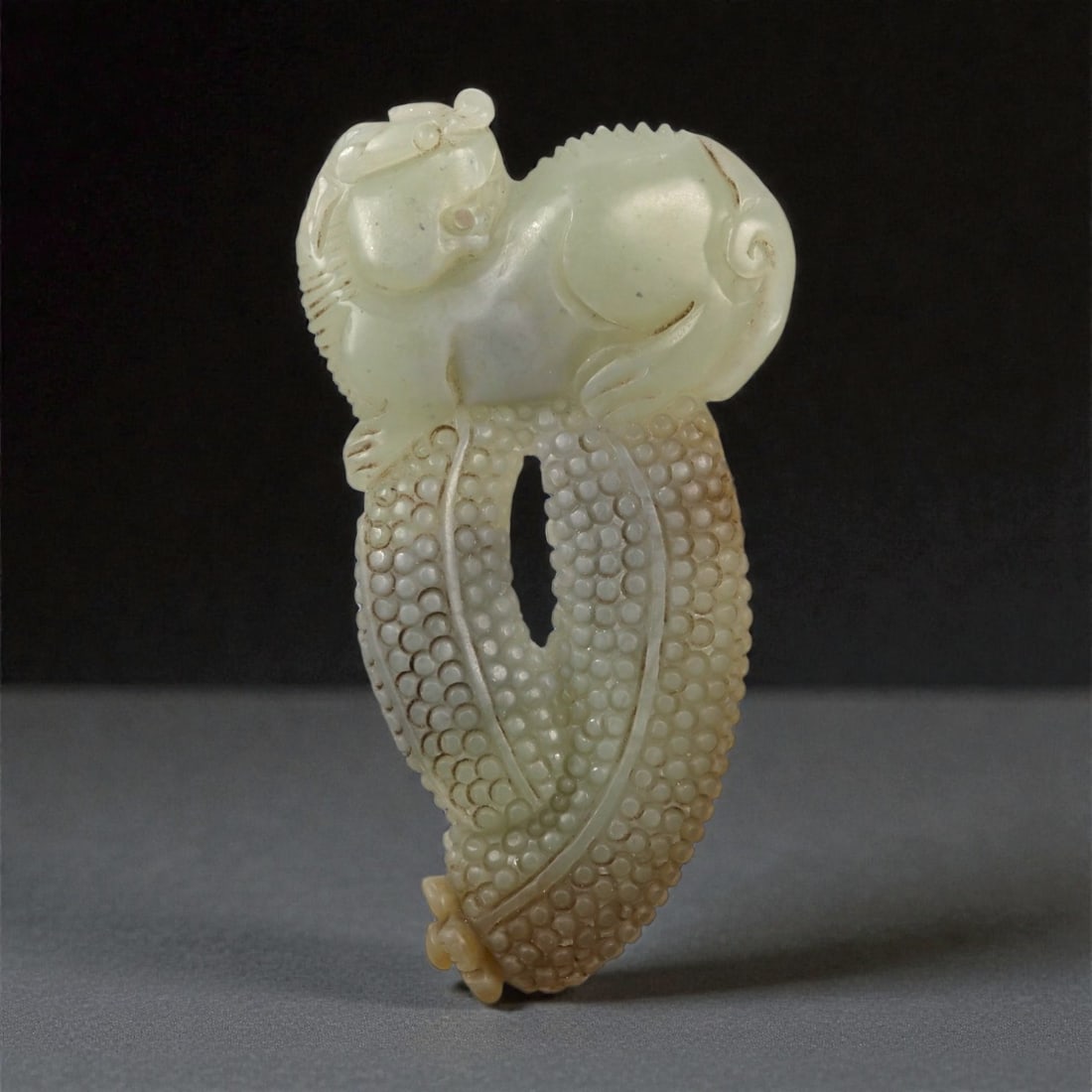 A Chinese Nephrite Jade Pixie and Fruit Pendant, 20th Century, 54FA: Carved from pale celadon nephrite jade with areas of soft translucency and light russet inclusions, the object is composed as a vertical pendant combining a pixie (mythical winged beast) above a styli