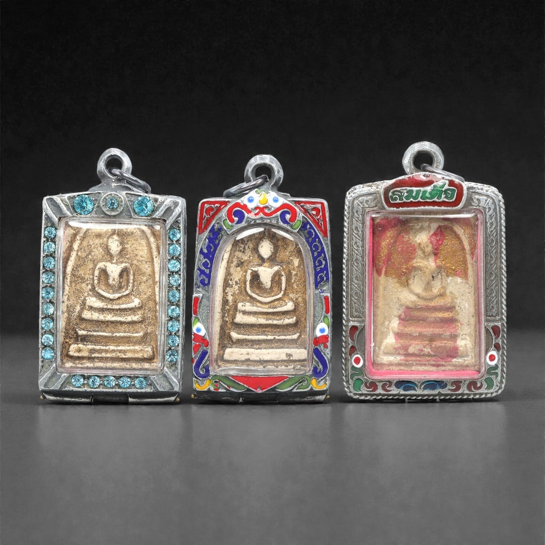 A Group of Three Thai Terracotta Buddha Amulets, 20th Century, 80XJ: Each amulet is formed from terracotta, enclosed in a silver-toned metal case with enamel and glass accents, and fitted with a suspension loop for wear. The amulets depict a seated Buddha in bhumispars